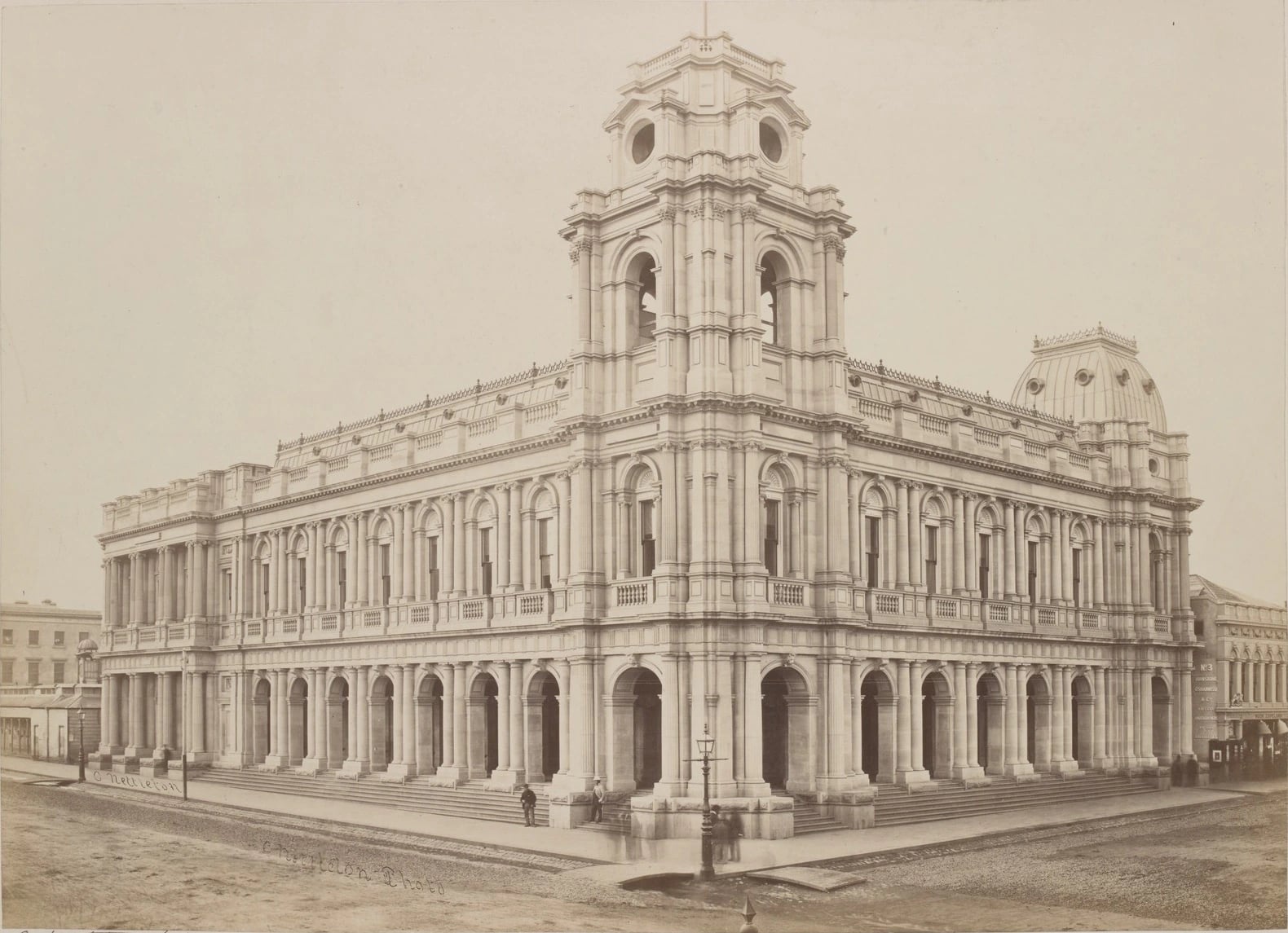 An 1868 photograph of Melbourne's General Post Office: a two-storey stone building on a street corner, a long arcade of round arches at pavement level, a square tower rising over the corner, and a few figures standing on the empty roadway.