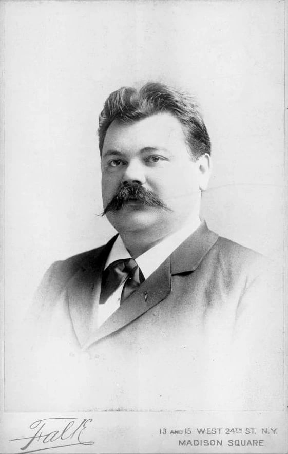 A black and white studio photograph of the violinist Adolph Brodsky, head and shoulders, a heavy-set man in his forties with a broad moustache, dark swept-back hair and a pale suit with a dark tie. The photographer's signature and a Madison Square, New York address are printed on the mount below.