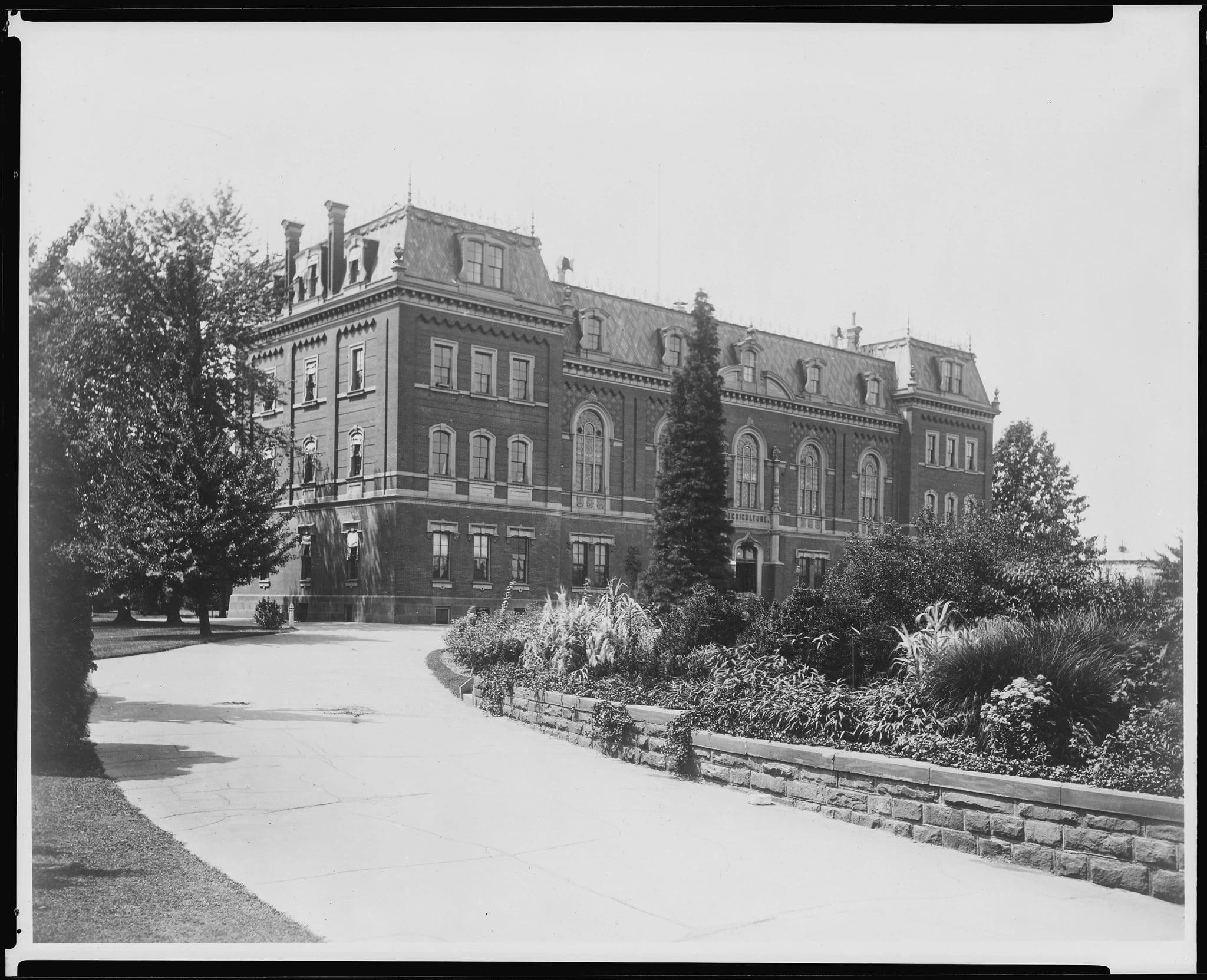 A black-and-white photograph of a large Victorian brick building with a mansard roof and arched windows, seen across a curving path with ornamental planting and trees in front of it.