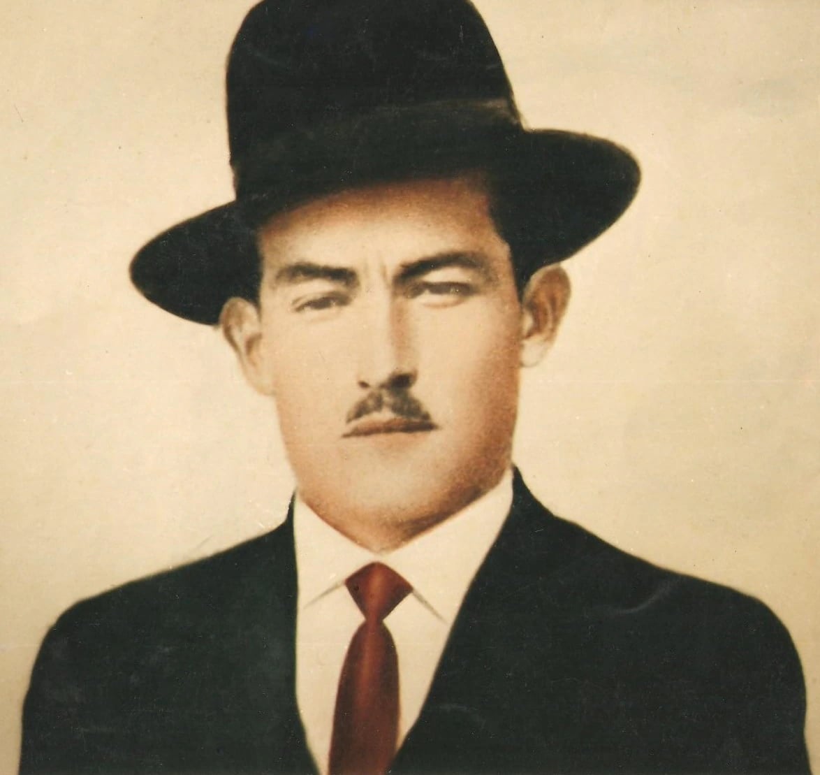 A tinted head and shoulders portrait photograph of a man with dark hair, a heavy moustache and level dark eyes, wearing a black bowler hat, a dark jacket, a white collar and a red tie, against a plain cream background.