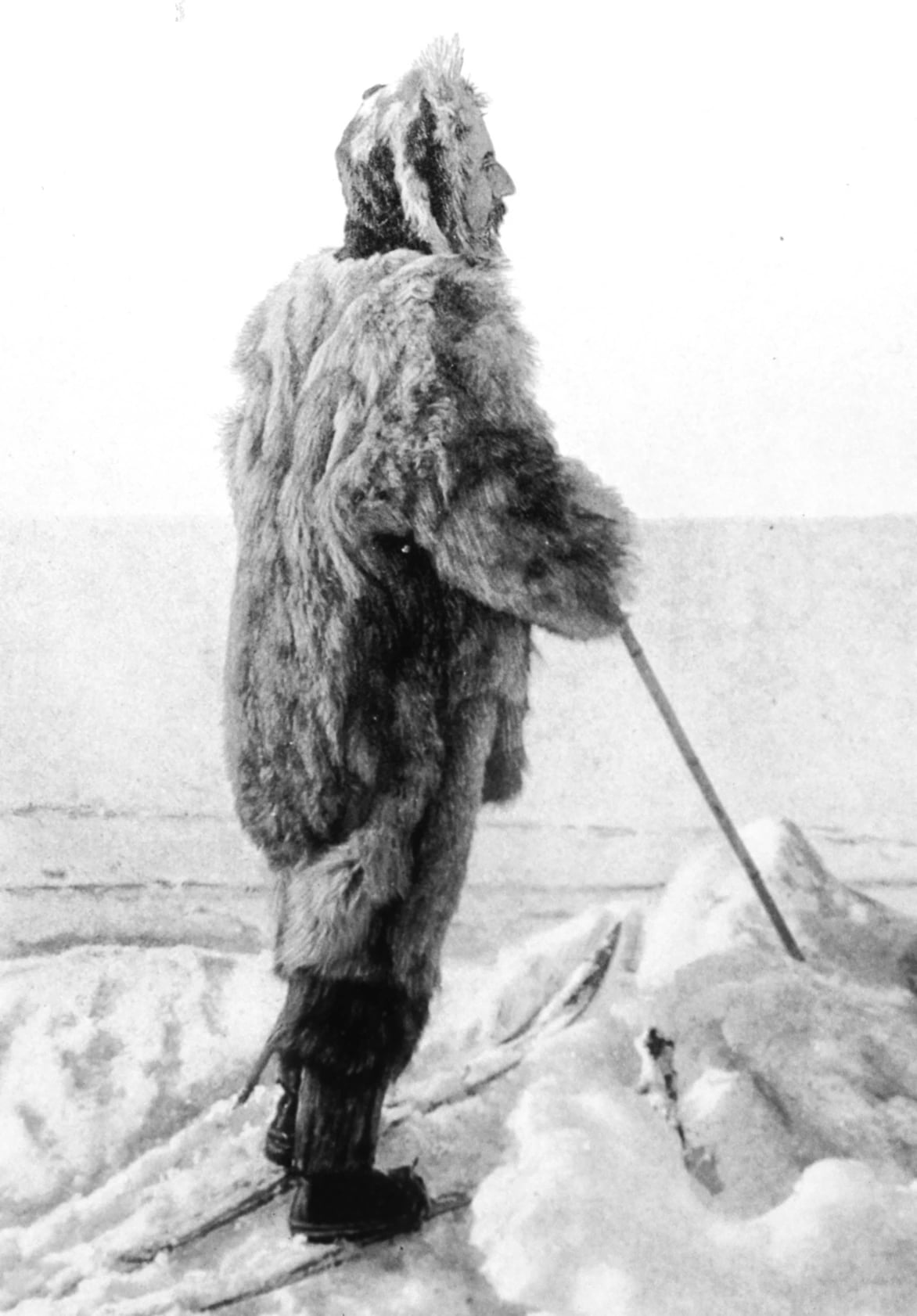 A black and white photograph of a bearded man in a full-length loose fur suit and fur hood, standing side-on on skis on broken ice, holding a single long ski pole.