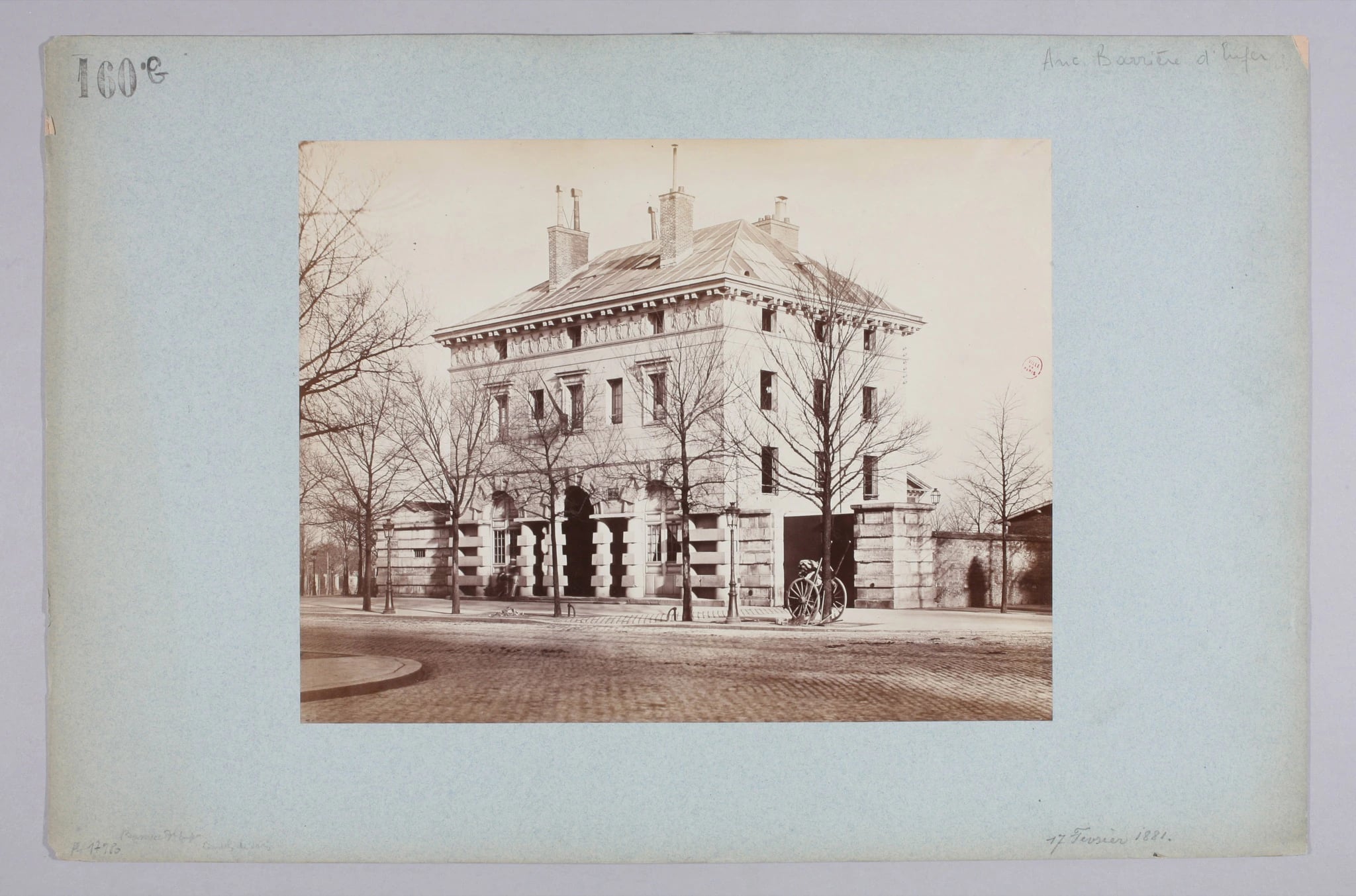 A sepia albumen photograph mounted on blue-grey card: a square neoclassical stone pavilion of two storeys, its upper wall carved with a frieze of panels and its roof hipped, flanked by low rusticated gate walls, with bare winter trees in front of it and a bicycle leaning against a gatepost on an empty cobbled street.