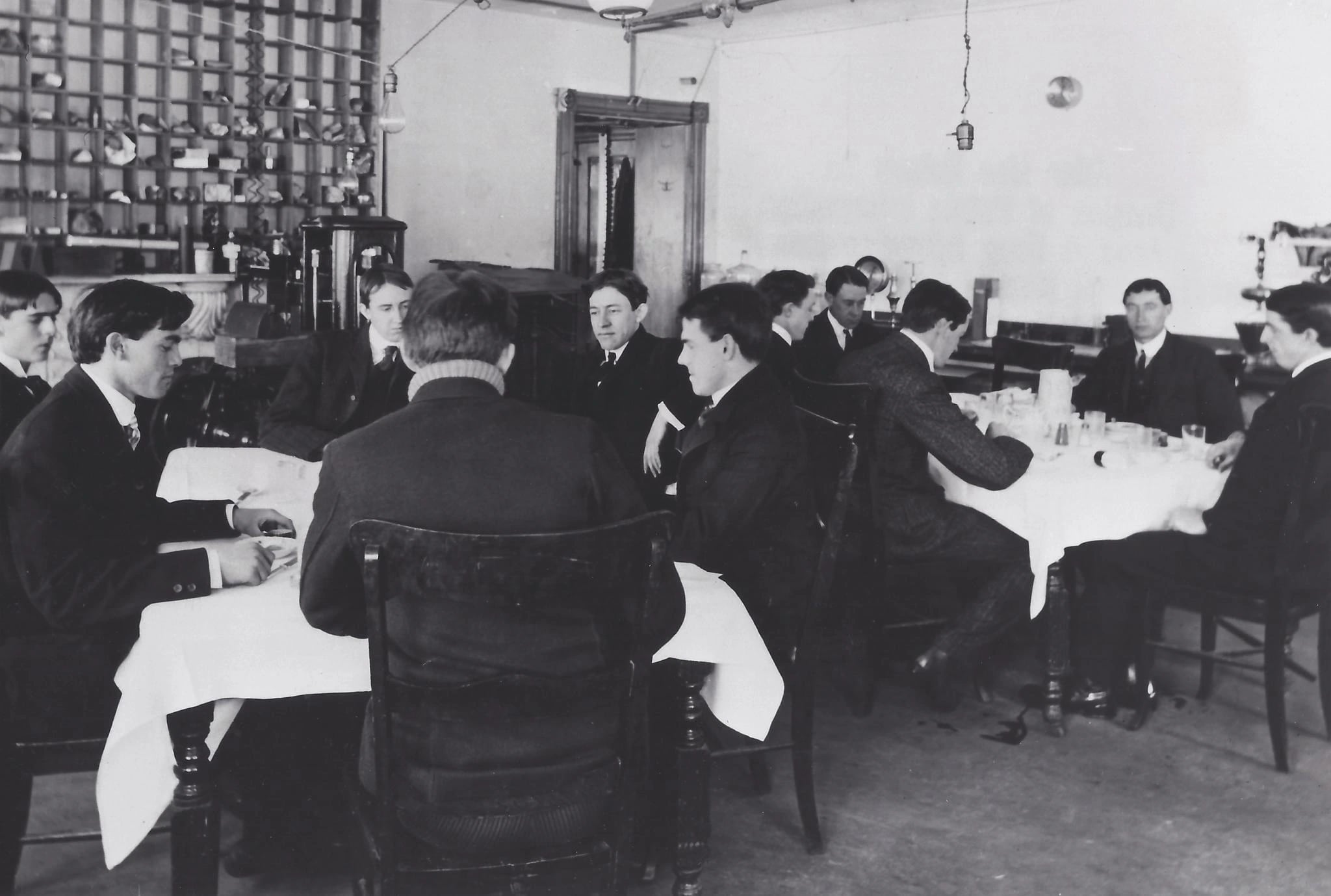 A black-and-white photograph of a plain room with two tables laid with white cloths, a dozen young men seated at them in suits, bare electric bulbs hanging on flexes, and a wall of pigeonhole racks holding specimen jars and tins at the left.