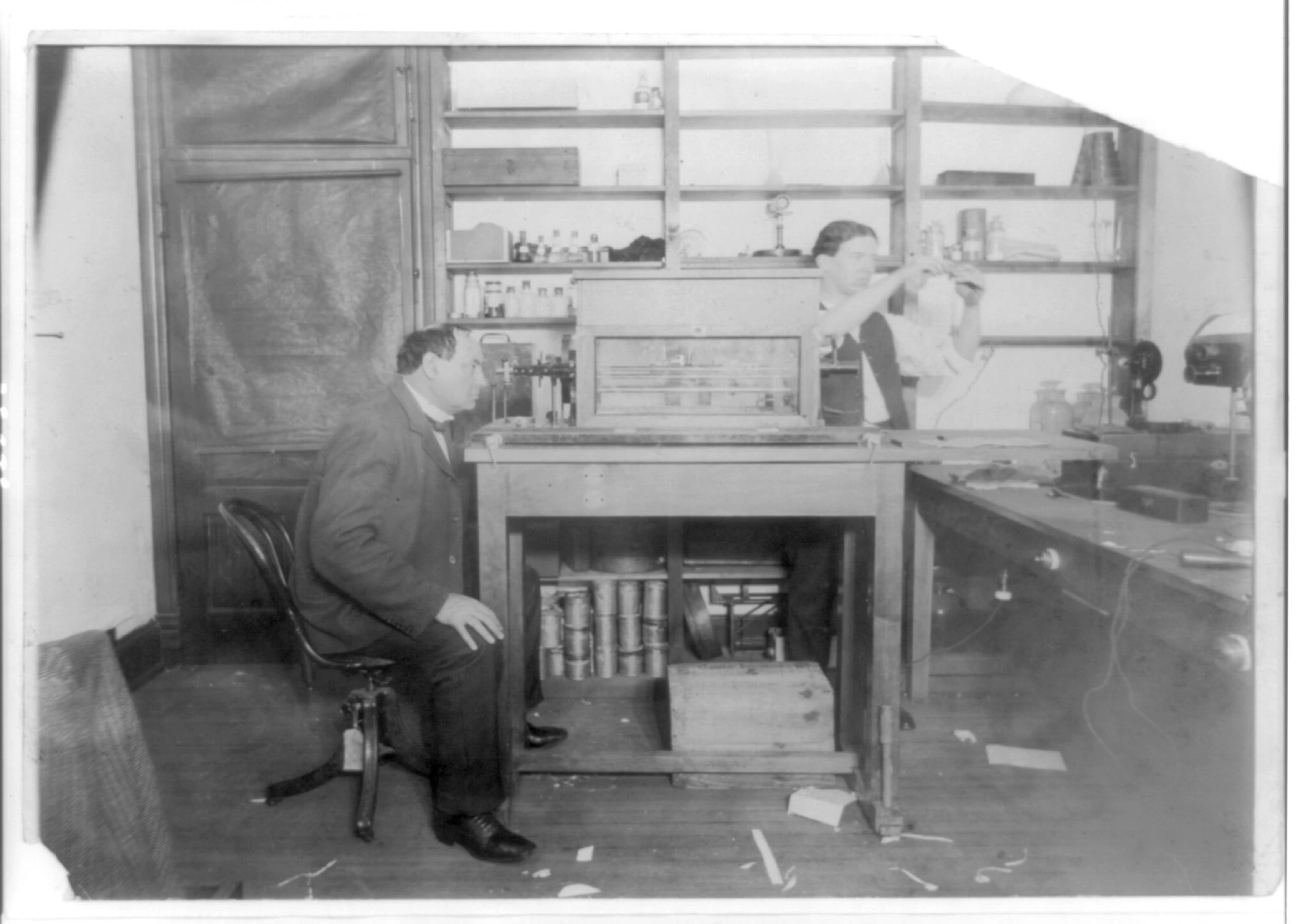 A black-and-white photograph of a laboratory: a moustached man in a dark suit sits on a swivel chair at the left, a second figure works at an apparatus on a wooden bench, shelves of bottles line the wall behind, and rows of tin cans are stacked on a shelf under the bench.