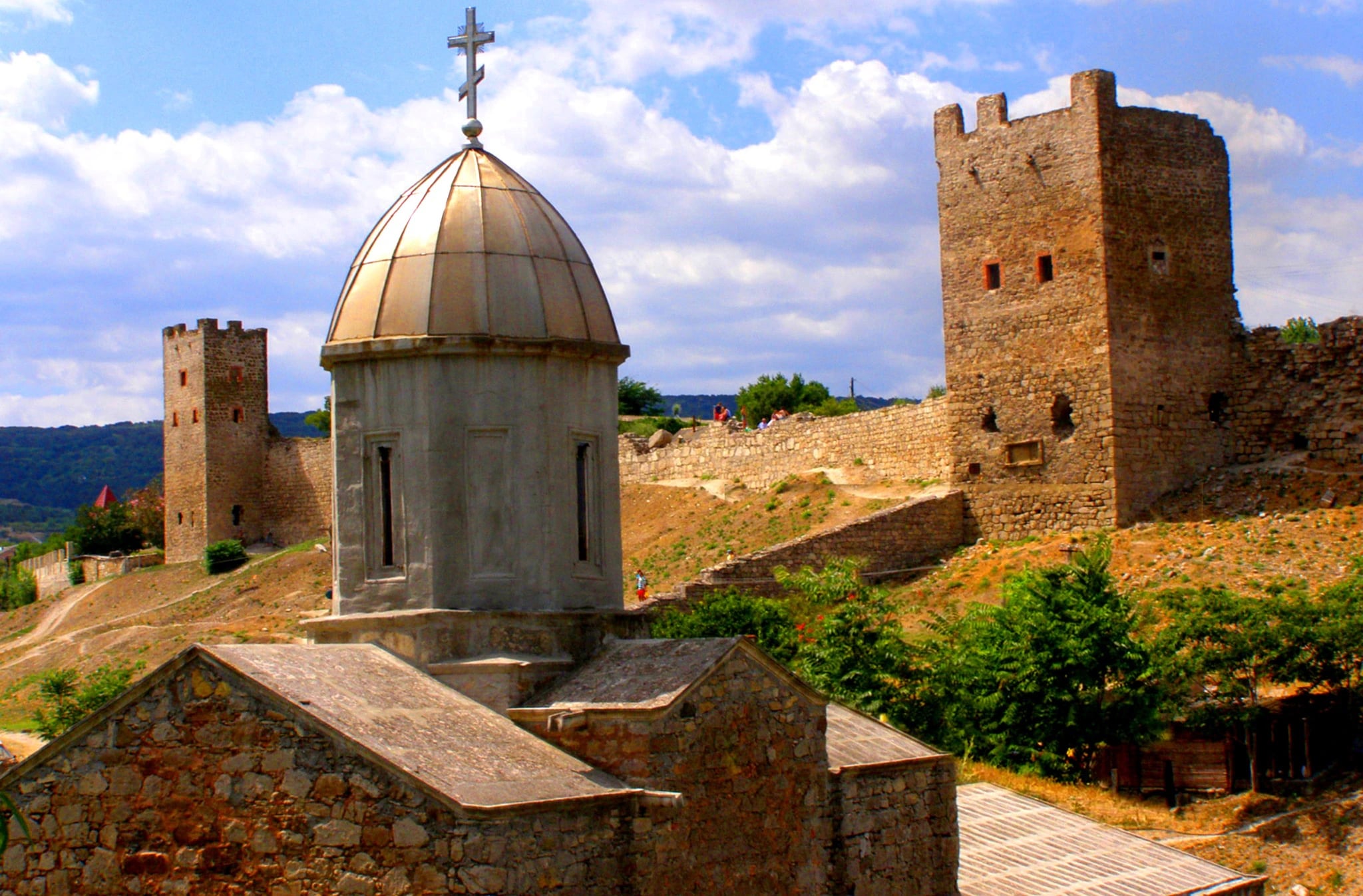 A photograph of two ruined medieval stone towers standing on dry open hillside above a small domed church with a metal cross. A crenellated curtain wall runs between them across the slope, with trees and scrub below and a bright blue sky with white cloud behind.