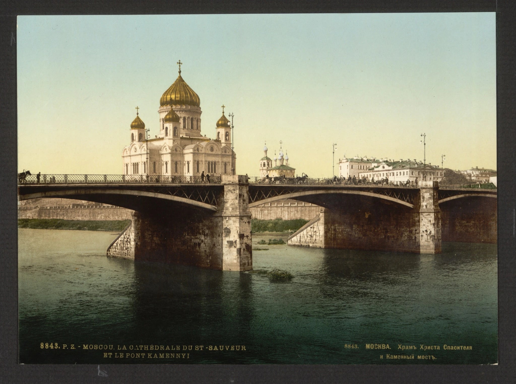 A colour photochrom print of a large white cathedral with a great gilded central dome and four smaller gilded domes, seen across a river from beyond a long stone bridge. Carriages and figures cross the bridge, low buildings and a smaller church stand along the far bank, and a bilingual French and Russian caption is printed along the bottom of the mount.