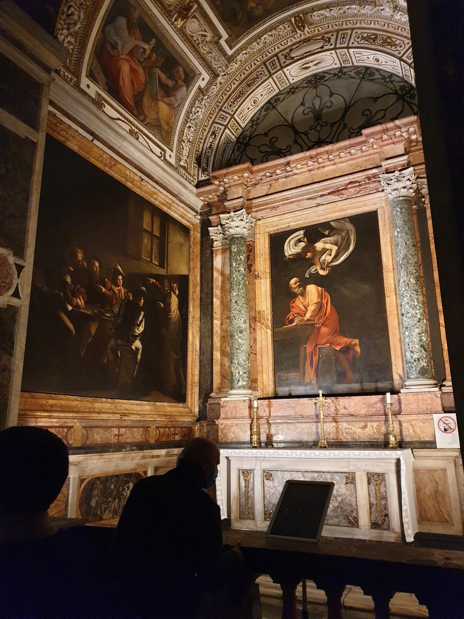 The Contarelli Chapel photographed from the nave in low light: The Calling of Saint Matthew on the left wall, The Inspiration of Saint Matthew in a marble frame over the altar, frescoes in the vault above, and two visitors standing in the dark in front of the rail.