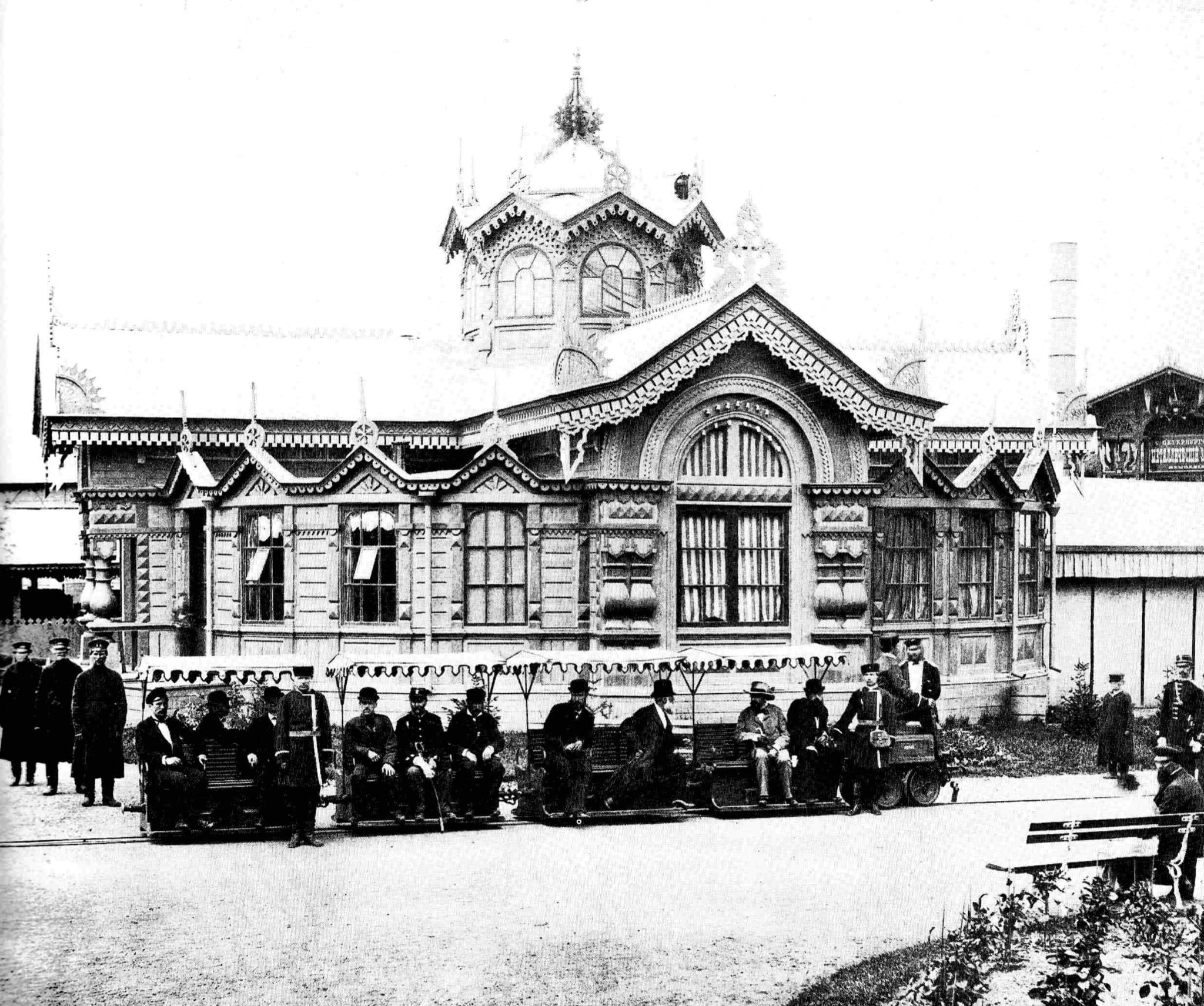 A black and white photograph of a short electric train standing on rails outside a wooden exhibition pavilion. Two open cars with scalloped canopies carry about a dozen men in top hats and bowlers on bench seats, with a small four-wheeled locomotive at the far end. Behind them rises an elaborately carved timber building with fretwork gables, arched windows and a small cupola, hung with bunting. Uniformed attendants stand at either side.
