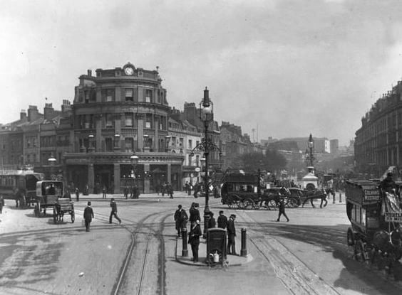 An early twentieth-century photograph of the Elephant and Castle junction: a domed corner public house with a clock, tram rails curving across the roadway, horse vans, an ornate lamp standard and pedestrians.