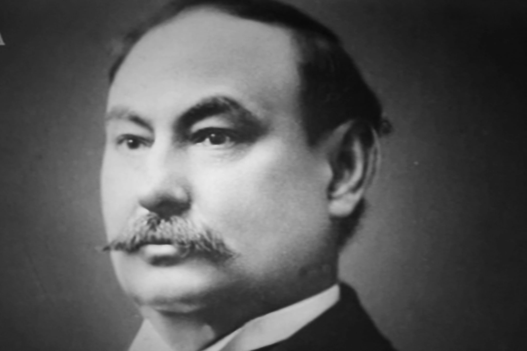 A studio photograph of a middle-aged man in three-quarter profile against a dark background: heavy brows, a broad moustache, dark hair swept back, wearing a dark jacket and white collar.