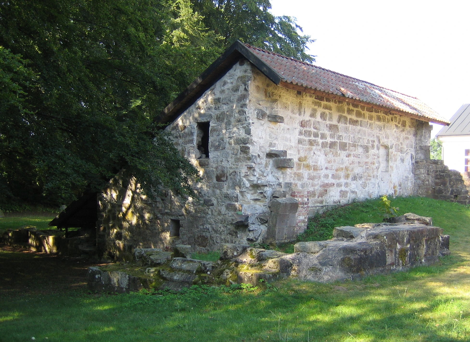 A small medieval stone building with a red tiled roof and rough whitewashed walls stands on a lawn in summer sunlight, a large tree leaning over it from the left. In the grass in front, low courses of excavated stone foundation run across the picture.