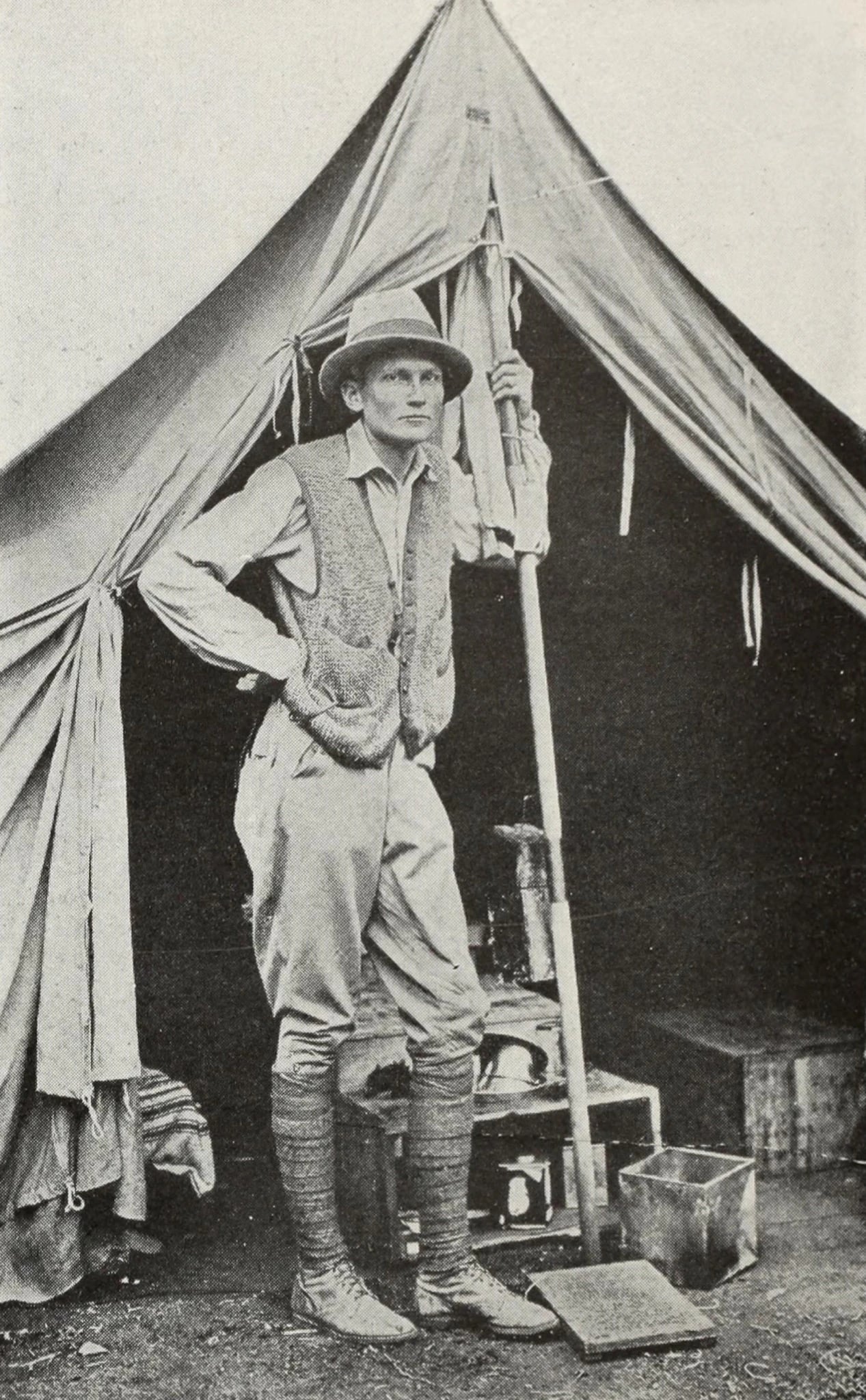 A black and white photograph of a young man standing at the open flap of a canvas ridge tent. He wears a soft brimmed hat, a knitted waistcoat over a shirt, riding breeches, tall laced boots and puttees, one hand on his hip and the other holding the tent pole. Packing cases and a metal box stand on the ground beside him.