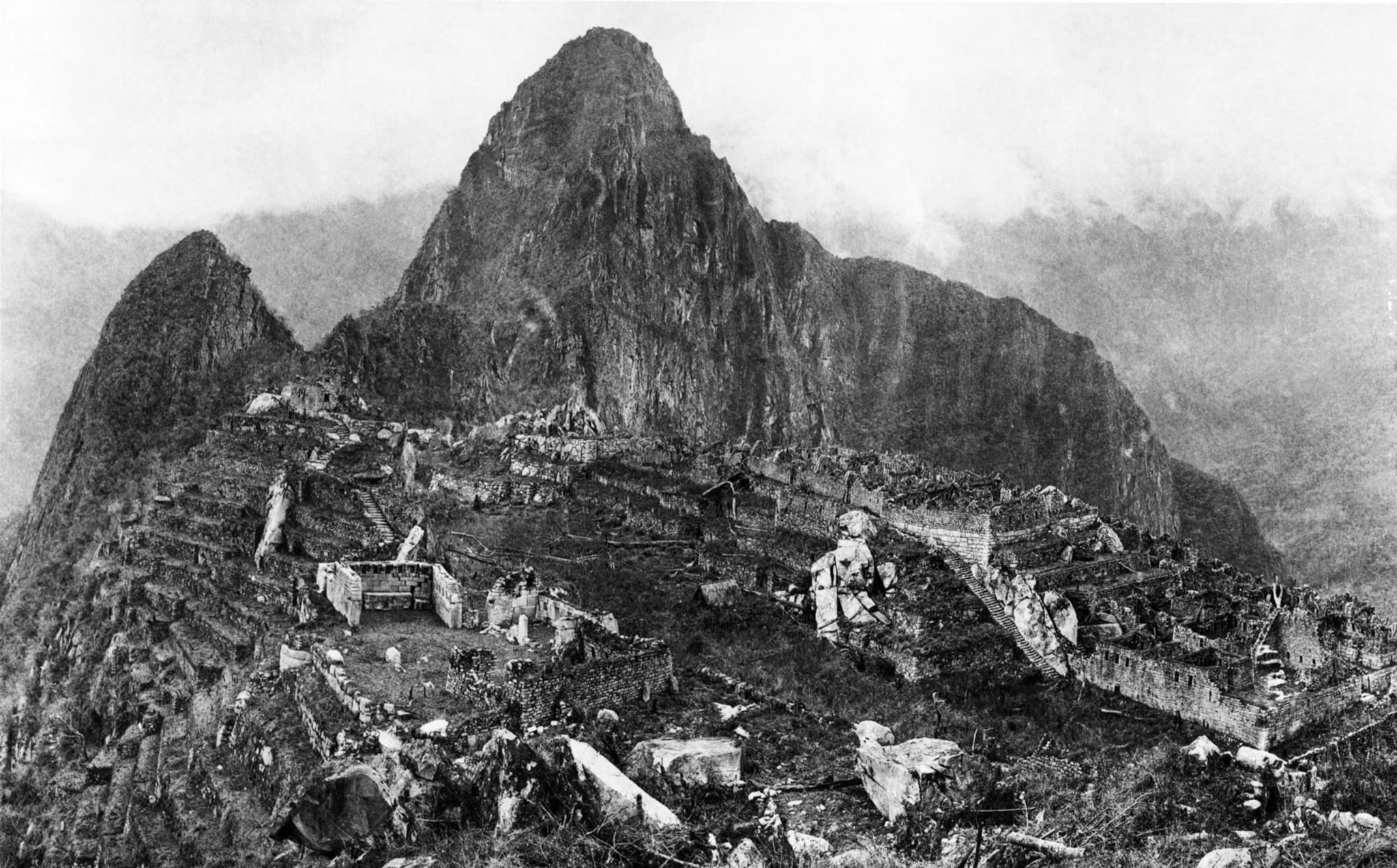 A black and white photograph looking across the cleared ruins of Machu Picchu, with stone walls, plazas, staircases and terraces laid bare on the saddle of the ridge, and the steep sugarloaf peak of Huayna Picchu rising directly behind them.
