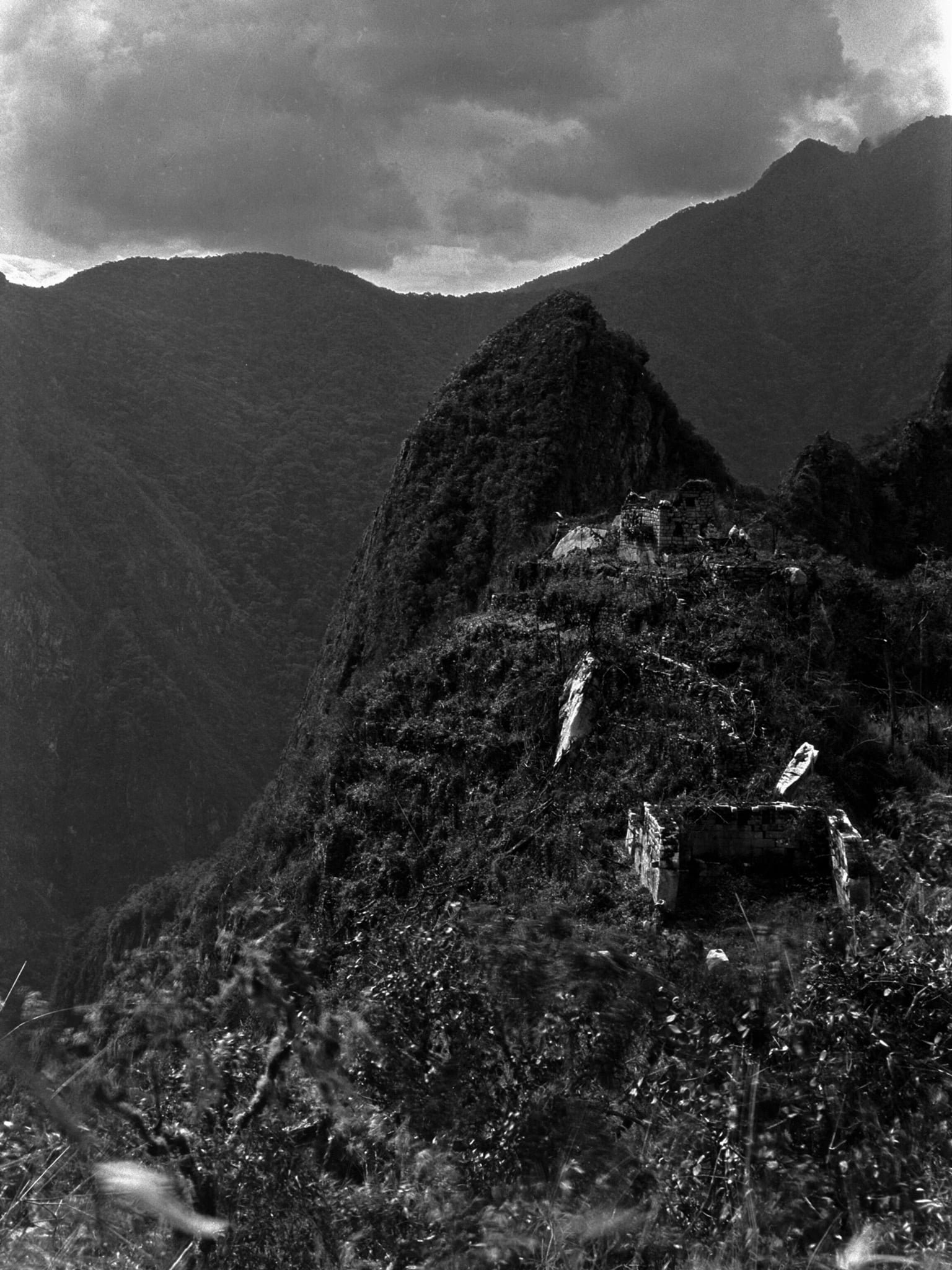 A black and white photograph of a steep forested ridge below a dark peak, with cloud-filled valleys falling away on either side. Stone buildings and terrace walls show faintly through dense vegetation along the crest and on the slope below it, almost lost in the scrub.