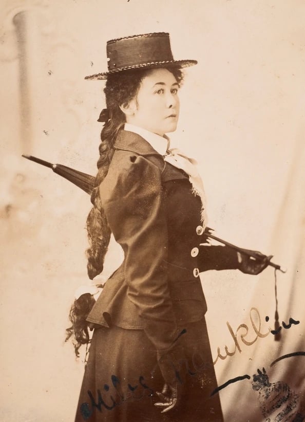 A sepia studio portrait of a young woman in her early twenties, seated and turned slightly to one side, in a high-necked pale blouse with a dark bodice. Her hair is dressed up and back from her face, and she looks away from the camera with a composed, level expression.