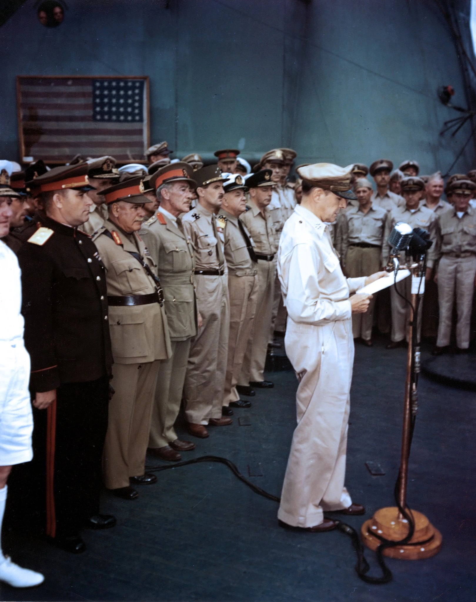 A colour photograph of the surrender ceremony aboard a warship: General Douglas MacArthur in plain khaki reads from a sheet of paper at a microphone stand, a row of Allied officers in uniform standing behind him, and a framed American flag mounted high on the grey bulkhead at the upper left.