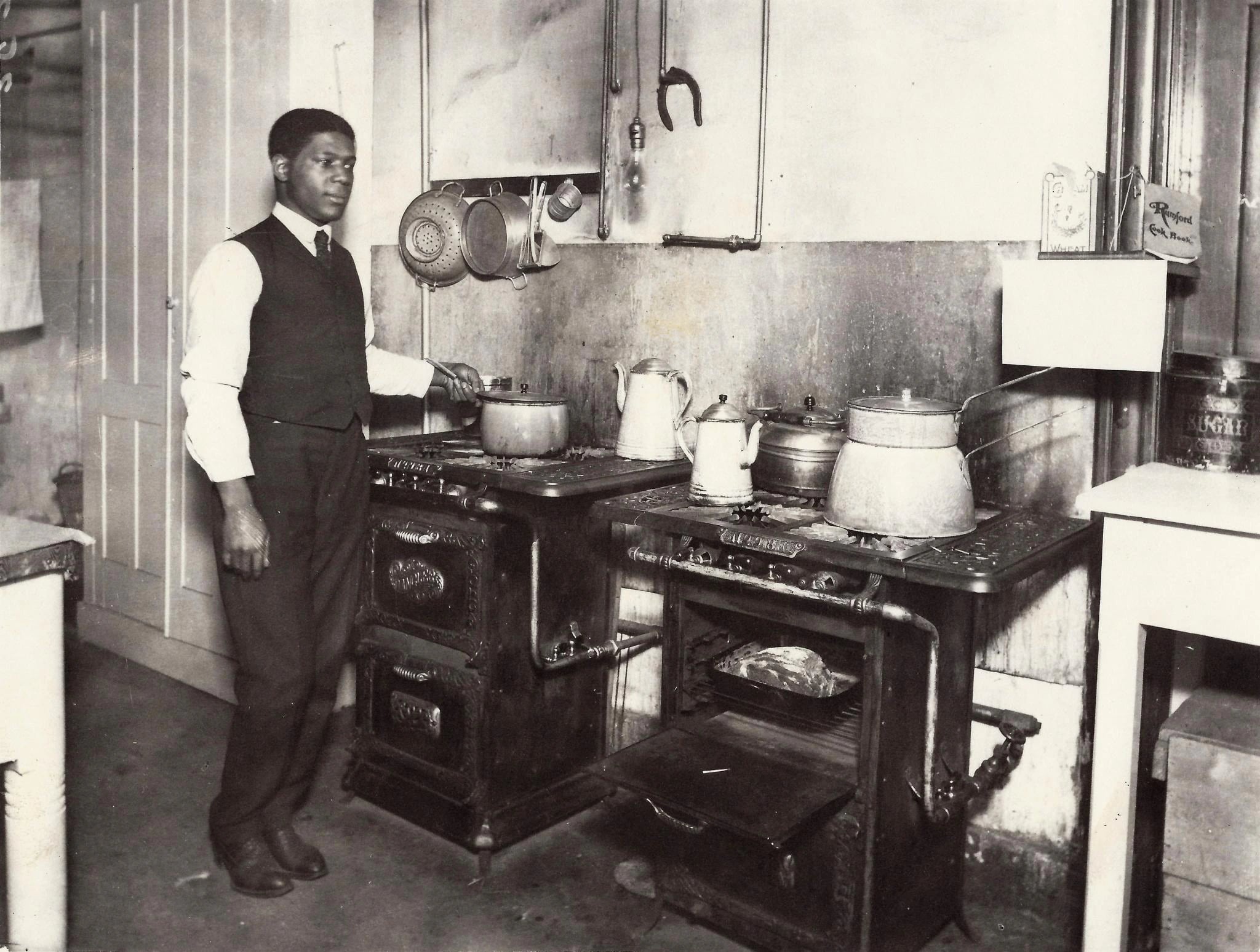 A black-and-white photograph of a young Black man in a waistcoat and shirtsleeves standing beside two cast-iron gas ranges in a basement kitchen, one hand on a pan, with kettles and covered pots on the hobs and a joint of meat visible in an open oven.