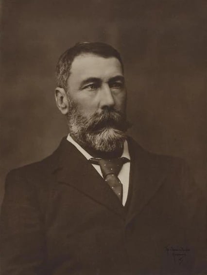 A black and white studio portrait of a middle-aged man in a dark three-piece suit and high stiff collar, seated and facing slightly left. He has short grey hair, a full moustache and a serious, heavy-featured face.
