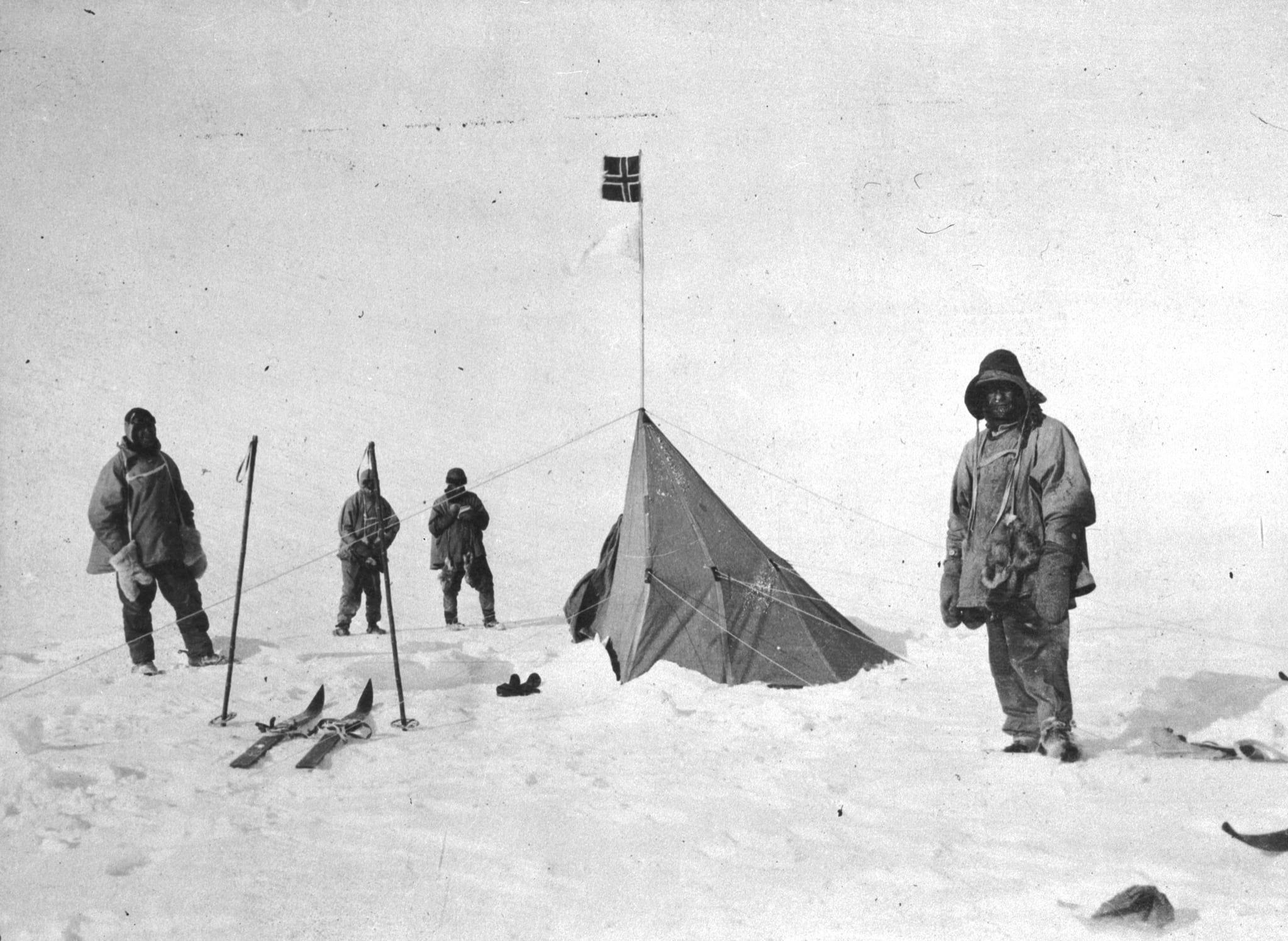 A black and white photograph of four men in windproof clothing standing around a small pyramidal tent flying a Norwegian flag, with skis and ski poles planted in the snow beside them.