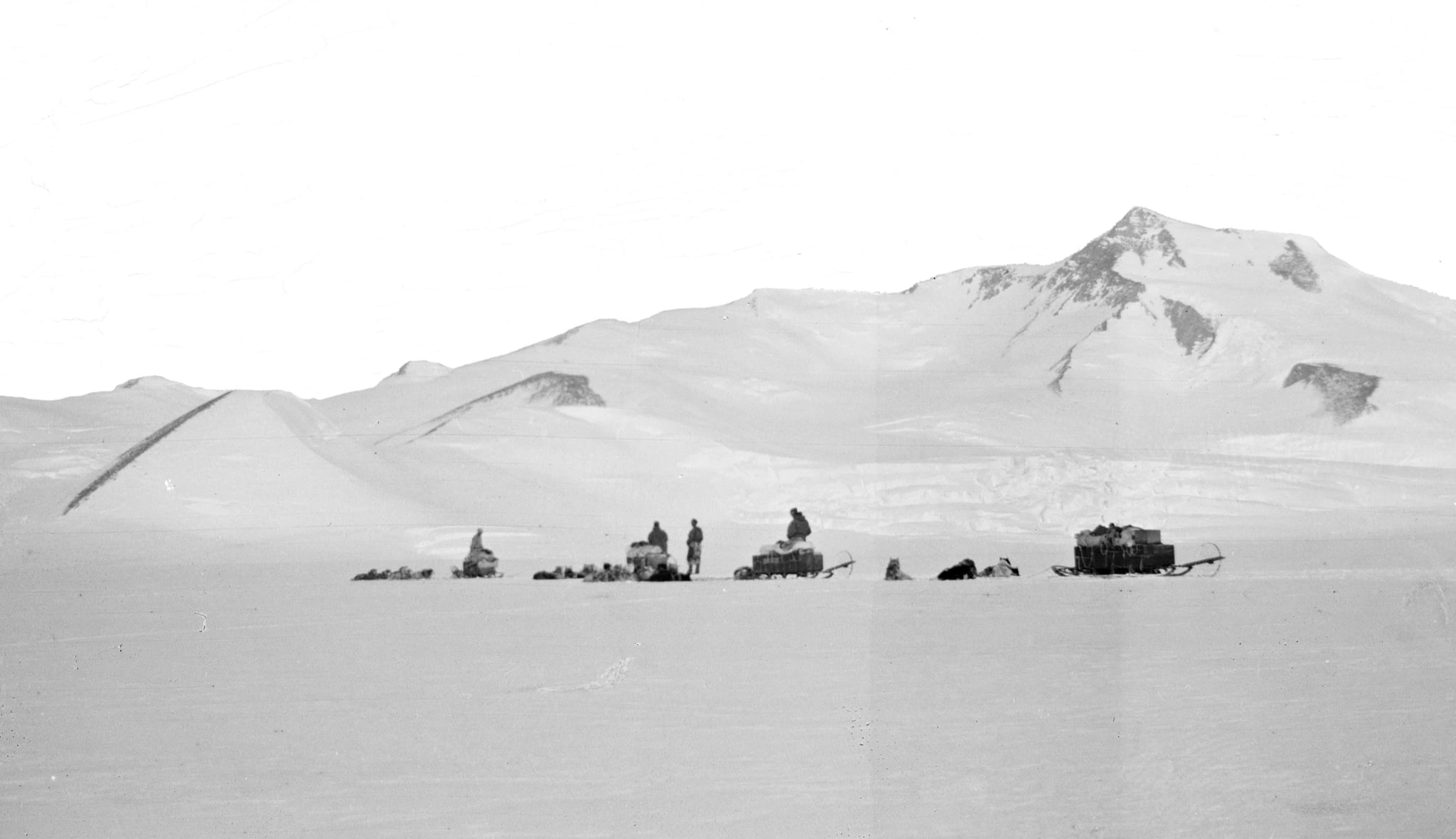 A black and white photograph of several loaded sledges and dog teams strung out across a vast snowfield, with a dark rocky mountain ridge rising behind them.