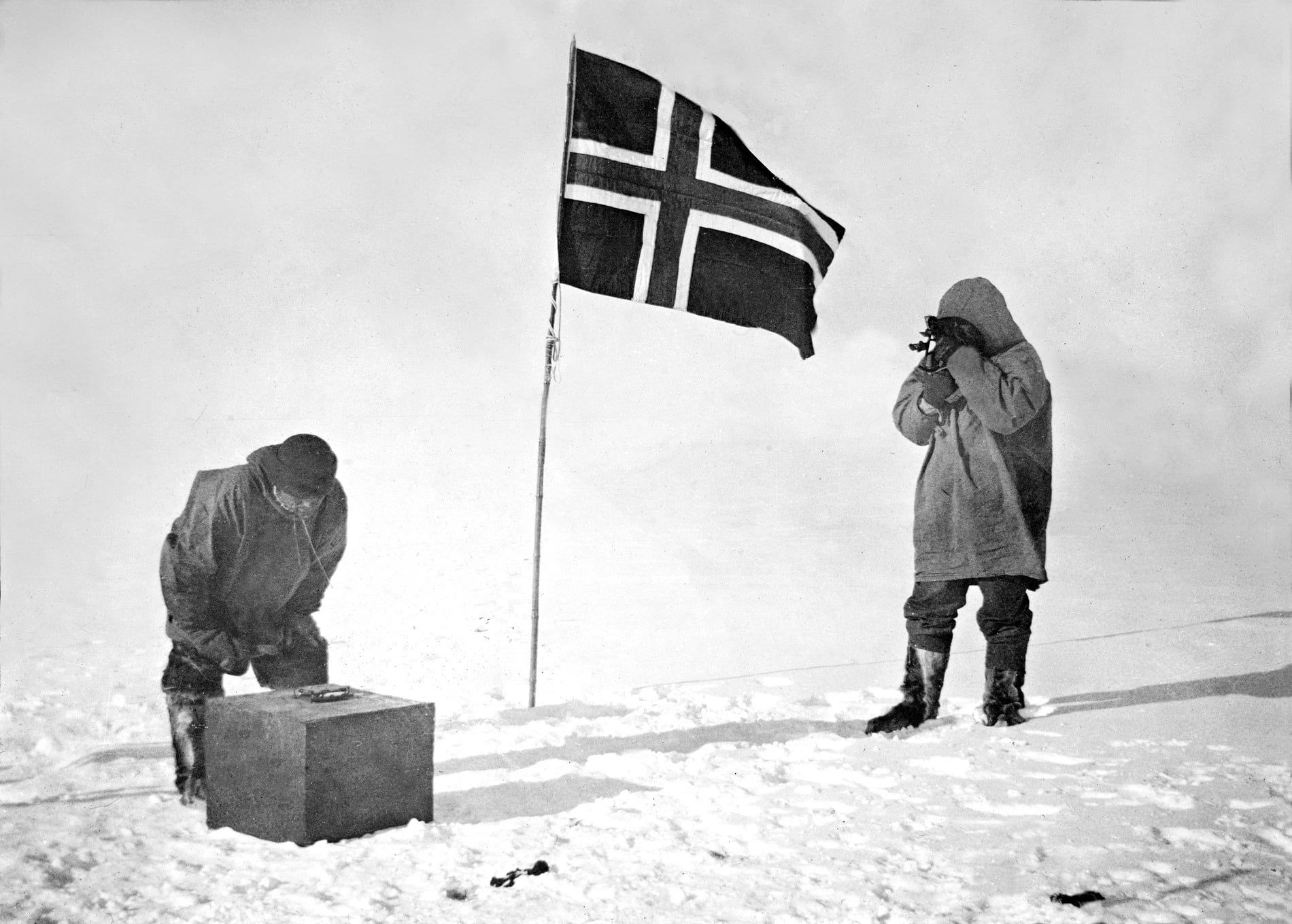 A black and white photograph of two men in fur clothing on snow beside a Norwegian flag on a pole: one stands taking a sight through a hand-held instrument, the other bends over a box on the ground.