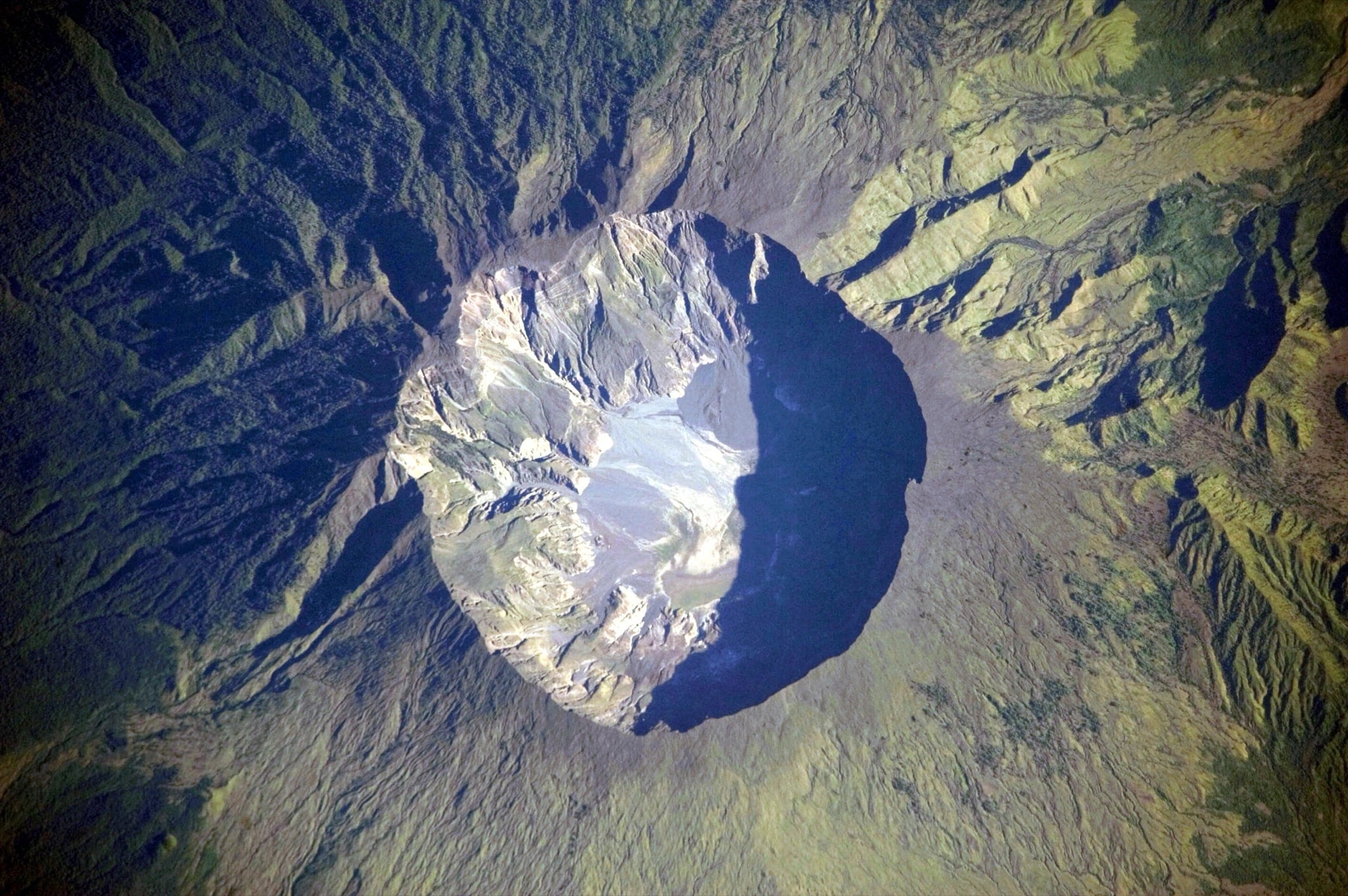 An astronaut photograph looking straight down on a volcanic summit: a near-circular crater several kilometres wide, its western rim catching the sun in pale grey and its eastern half filled with deep black shadow, sitting in a ridged cone of dark green forest that runs out to open pale ground at the edges of the frame.