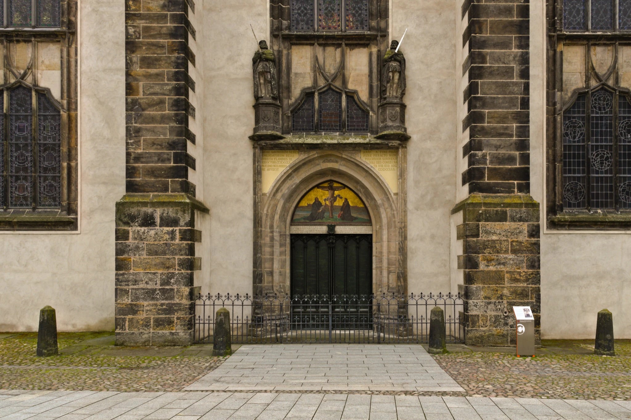 A pointed stone portal in the pale plastered wall of a church, flanked by two heavy stone buttresses and by tall traceried windows. Dark bronze double doors covered in columns of small lettering stand deep in the arch, under a painted tympanum of the Crucifixion and a band of gilt Latin inscription. Carved standing figures sit on brackets either side, a low iron railing crosses the front, and cobbles and paving run up to it.