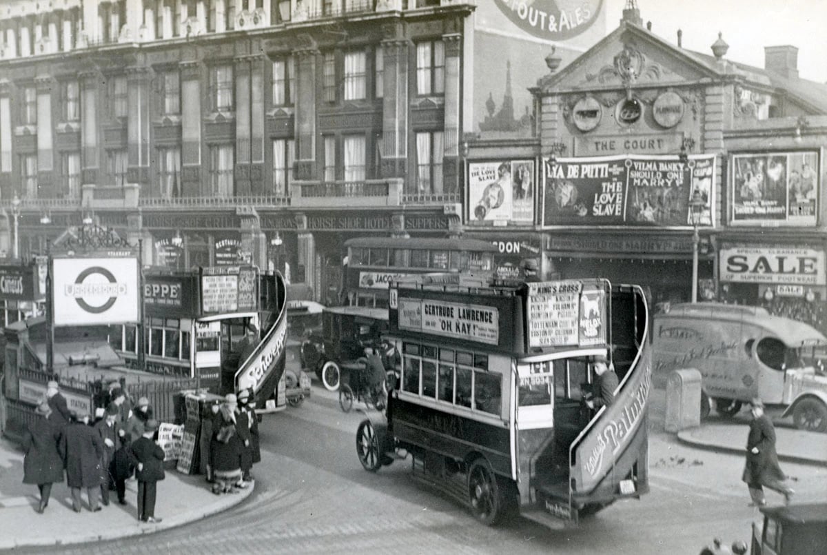 A 1927 photograph of Tottenham Court Road in London: an open-topped double-deck bus, a cinema hung with film posters, shop windows advertising a sale, and people in winter coats on the pavement.