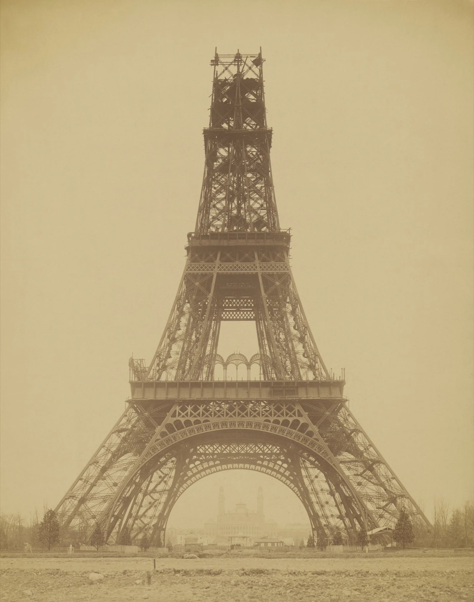 A sepia albumen photograph of the Eiffel Tower unfinished, seen head-on from ground level across an expanse of bare rubble-strewn earth. The four curving legs and the first two platforms are complete, and above them a narrower, scaffolded section of open lattice tapers up and stops short against a blank pale sky. Framed inside the great arch between the legs, small in the distance, stands a long domed palace with two tall towers.